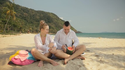 The cheerful love couple holding and eating slices of watermelon on tropical sand beach sea. Romantic lovers two people caucasian spend summer weekend in outdoor. Hat, backpack white shirt beachwear.