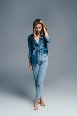 young woman in denim shirt and jeans touching hair while standing barefoot on grey