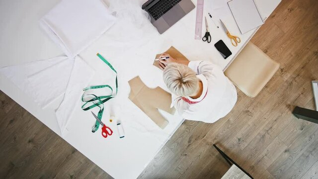 Female, tailor of sewing workshop is drawing pen around pattern which laying on white fabric while standing at table