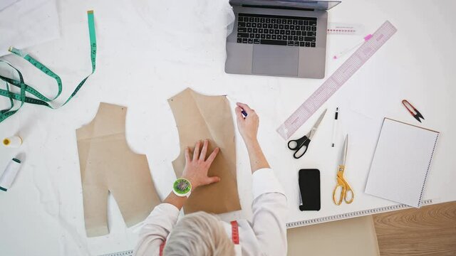 Woman tailor of sewing workshop is drawing pen around pattern laying on white fabric, standing at table