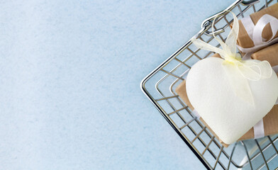 White heart decorated by bow lies with gift boxes in shopping cart above light blue background
