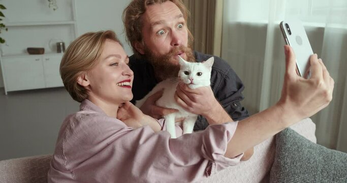 Two people caucasian family couple bearded man boyfriend husband and blonde beautiful wife girl sitting together on sofa hugging white beloved cat animal pet waving hand and paw at smartphone camera