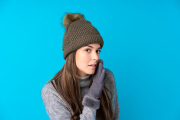 Teenager girl with winter hat over isolated blue background whispering something