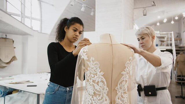 Females tailors are working on wedding dress hanging on mannequin, measuring and pinning it up, standing at sewing workshop