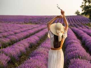 A young woman with long hair in a straw hat and dress stands in a lavender field. Photo from the back.