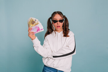 Fashionable girl with collected hair wearing black sunglasses and white pullover posing with a wad...