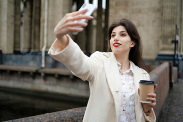 Beautiful girl makes a selfie on the phone. With coffee in hand