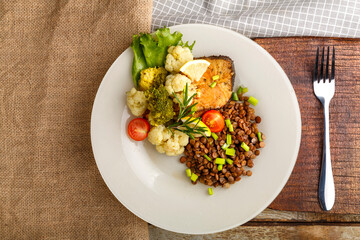 Baked fish with broccoli and lentils in a white plate with rosemary and lemon on a napkin and a round stand next to a fork.