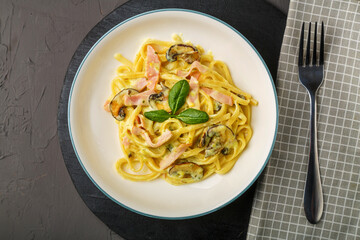 a plate of pasta with ham and mushrooms in a creamy sauce with basil on a black round stand with a fork on a napkin.