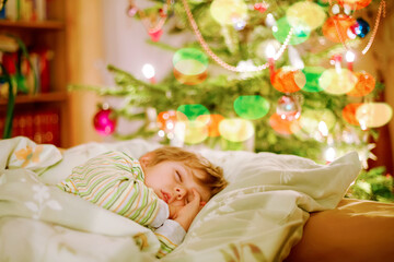 Obraz na płótnie Canvas Little cute blond boy sleeping under Christmas tree and dreaming of Santa at home, indoors. Traditional Christian festival. Happy kid child waiting for gifts on xmas. Cozy soft light