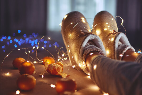 Ripe Mandarins Orange, Warm White Christmas Lights Garland And Female Legs Wearing Warm Fluffy Soft Winter Slippers At Cozy Home At Christmas Eve