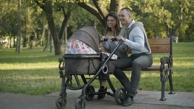 Wide shot of happy loving couple of young parents sitting with baby carriage in summer park and talking. Smiling Caucasian man and woman enjoying sunny evening outdoors with infant in baby stroller.