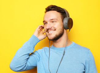 Handsome man wearing blue sweater and headphones and holding mobile phone over yellow background