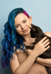 Obraz premium Girl with creative blue coloring and a rainbow in her hair on a blue background holds black cat. Modern minimalistic bright photography for advertising and social networks