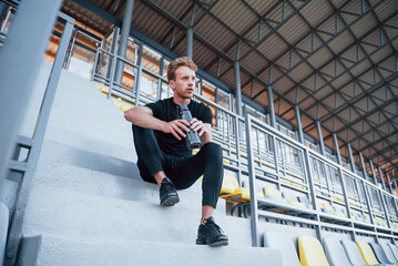 Sits with bottle of water. Sportive young guy in black shirt and pants outdoors at daytime