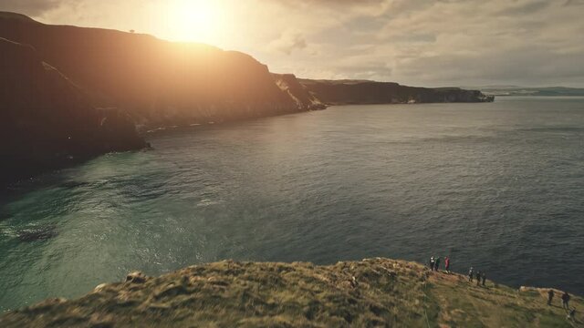 Aerial people on sun ocean shore. Tourists at sea bay with cliff coast. Nature seascape at Irish landmark. Rock isle landscape with travelers. Carrick Island, Northern Ireland, United Kingdom, Europe