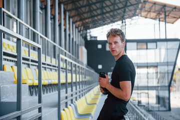 Fototapeta premium Standing and holding bottle of water. Sportive young guy in black shirt and pants outdoors at daytime