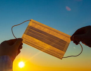 Protective medical face mask in rays of sunset orange sun and blue sky. Girl holds the mask with...