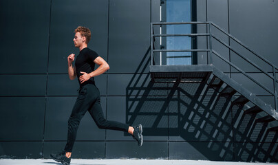 Fototapeta na wymiar Running near black building. Sportive young guy in black shirt and pants outdoors at daytime