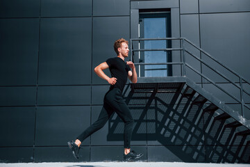 Fototapeta na wymiar Running near black building. Sportive young guy in black shirt and pants outdoors at daytime