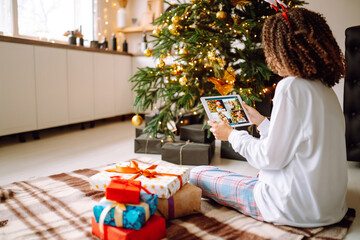Christmas online holiday. Happy woman having video call with their family or friends. Young woman uses a digital tablet near decorated festive tree at home. Virtual meeting. Covid-2019.