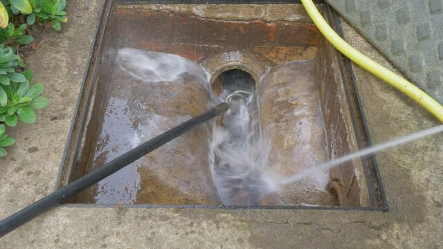 Closeup POV shot of a gloved hand holding a power hose while directing the water flow at  a drain, after clearing a pipe blockage with a flexible rod and worm screw.