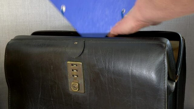 Closeup POV shot of a man&rsquo;s hands opening an old leather briefcase, placing a blue document folder insider, then shutting the case latch.