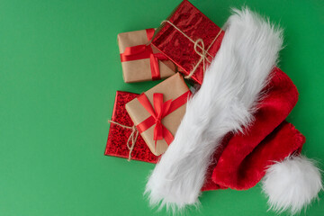 close up image of four wrapped gift boxes tired up with ribbons look out from big red santa hat on green background. Copy space. Holiday concept