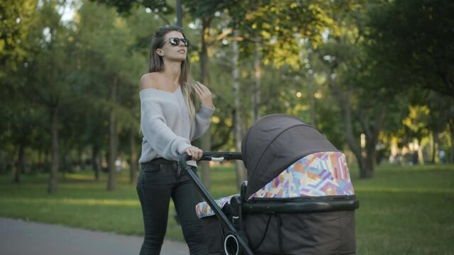 Gorgeous brunette Caucasian woman in sunglasses walking along the alley in summer park with baby stroller. Portrait of confident young mother strolling with infant in baby carriage outdoors.