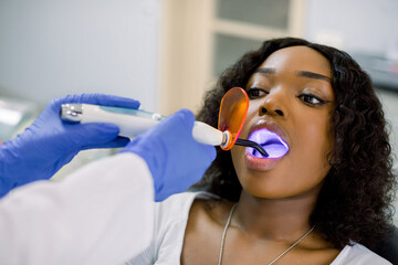 Close up of face of young African woman patient having dental treatment at dentist's office. Dentist using dental treatment with curing UV lamp. Tooth restoration with filling and polymerization lamp