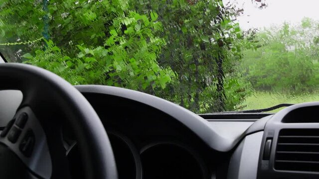 Rain flows over windshield glass surface shot from inside car with defocused foreground of black auto dashboard and steering wheel. Summer rainfall background for transport concept