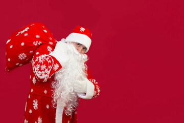 Santa holds a bag of gifts behind his back on a red background. © Roman Akimov