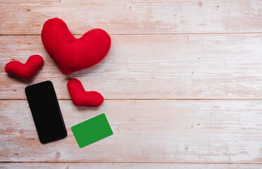 Three red toy soft hearts for handmade holiday. Credit chromakey card next to black smartphone on wooden table. Online shopping for Valentines day.