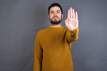Young handsome Caucasian man wearing yellow sweater against gray wall doing stop sing with palm of...