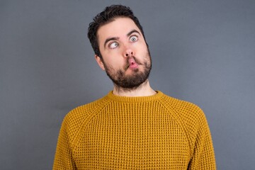 Obraz premium Young handsome Caucasian man wearing yellow sweater against gray wall making fish face with lips, crazy and comical gesture. Funny expression.
