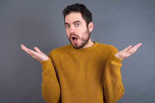 Frustrated Young handsome Caucasian man wearing yellow sweater against gray wall feels puzzled and hesitant, shrugs shoulders in bewilderment, keeps mouth widely opened, doesn't know what to do.