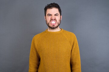 Young handsome Caucasian man wearing yellow sweater against gray wall keeps teeth clenched, frowns face in dissatisfaction, irritated because of much duties.