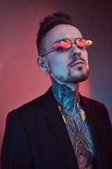 Fototapeta premium Portrait of a tattooed and fashionable hipster guy with stylish hairstyle in black suit weared with sunglasses poses in colourful background.