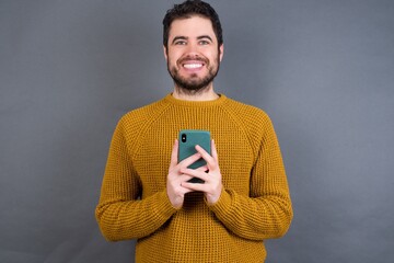 Young handsome Caucasian man wearing yellow sweater against gray wall enjoys distant communication, uses mobile phone, surfs fast unlimited internet, has pleasant smile, makes shopping online,