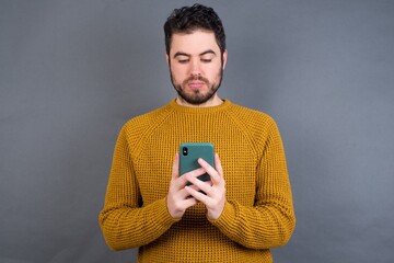 Excited Young handsome Caucasian man wearing yellow sweater against gray wall winking and eye hold smart phone use read social network news
