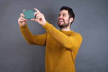 Young handsome Caucasian man wearing yellow sweater against gray wall taking a selfie to post it on...