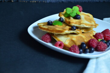 Homemade Belgium Waffles with fresh blueberries for breakfast. White plate, Viennese waffles with raspberries and mint. quick delicious breakfast on black background. top view copy space