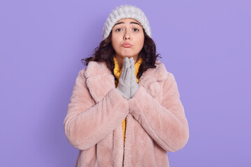 Attractive cute European girl in fur coat, cap and mittens, prays on lilac background, keeping...
