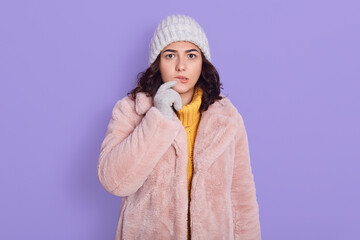 Pensive good looking young woman with dark hair looking thoughtfully at camera, dressed in fashionable pink coat and cap, looking concentrated, poses against blue background, keeping finger on lip.