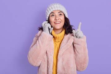 Happy young woman talking on mobile phone, having excited facial expression, pointing fore finger...