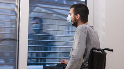 Disabled lonely man wearing face mask sitting in a wheelchair alone looking through window.