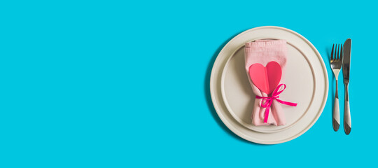 Banner with served table with plate and cutlery for the celebration of Valentine's Day. On the plate is napkin with paper heart. Flatlay website banner on bright blue background. Top view.