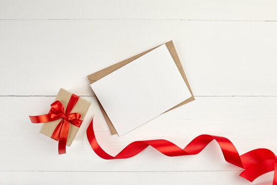 Greeting card mockup with paper envelope and red ribbon and gift box on white table