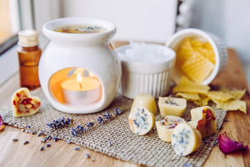 Fototapeta na wymiar Using homemade mini wax melts in aromatherapy lamp diffuser at home interior concept. Melts making ingredients on table for unbleached beeswax, solid coconut oil, essential oil, dried flowers.