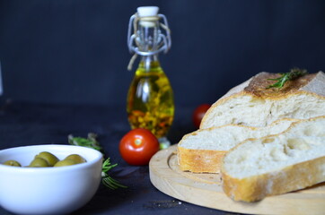 Italian ciabatta bread cut in slices on wooden chopping board with herbs, rosemary garlic and olives over dark grunge backdrop, top view